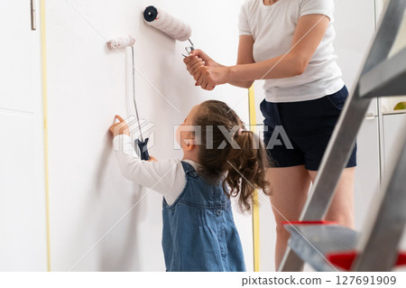 Little girl and her mother painting a wall together with rollers in the living room, family renovation concept. Little girl and her mother painting a wall together with rollers in the living room, family renovation concept. 127691909