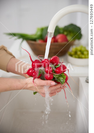 Close up view of a woman washes a bunch of red radishes under the pressure of flowing water in the white kitchen, a basket with tomato, broccoli and onions in the background, grapes Close up view of a woman washes a bunch of red radishes under the pressure of flowing water in the white kitchen, a basket with tomato, broccoli and onions in the background, grapes 127693449