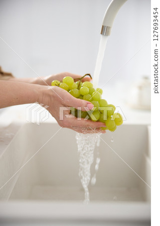 Close up view of a woman washes fresh green grapes under the pressure of flowing water in the white kitchen 127693454