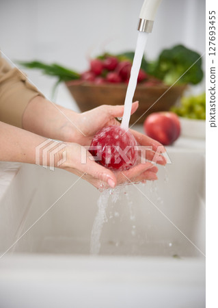 Close up view of a woman washes red peach under the pressure of flowing water in the white kitchen, a basket with radishes, broccoli and onions in the background, washed green grapes in a cup 127693455