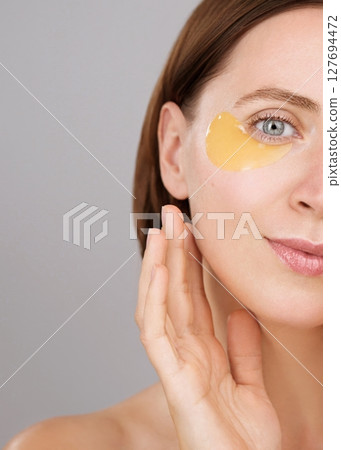 Half face. Eye patch ads concept. Aesthetic woman applying under-eye recovery patches. Skincare. Charming brunette woman in studio on grey background. Cosmetics Beauty routine Dermatology Rejuvenation Half face. Eye patch ads concept. Aesthetic woman applying under-eye recovery patches. Skincare. Charming brunette woman in studio on grey background. Cosmetics Beauty routine Dermatology Rejuvenation 127694472