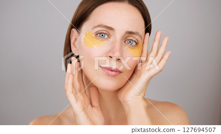 Eye patches, aesthetic woman applying under-eye recovery patches. Beautiful calm brunette woman posing in studio. Grey background. Cosmetics Skincare treatment Beauty routine Dermatology Rejuvenation 127694507