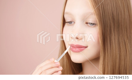 Cute young girl applying lip gloss. Caucasian blonde girl gen Alpha with blue eyes using make-up cosmetics, posing in studio on pink background. Concept of advertising cosmetics for teens. Copy space. 127694535