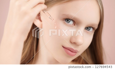 Skincare. Close-up portrait of a young Caucasian girl applying facial serum with a dropper. Pink background. Concept beauty ads, wellness blogs, cosmetic product promotions for teenagers, smooth skin 127694543