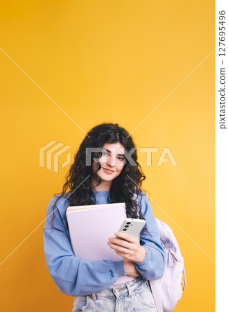 Smiling student using smartphone and holding notebook on yellow background Smiling student using smartphone and holding notebook on yellow background 127695496