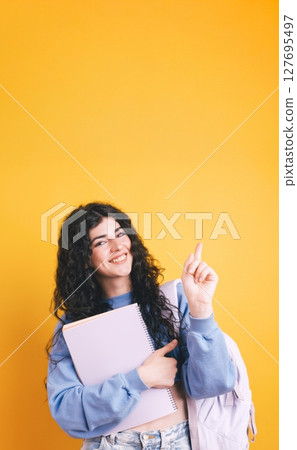 Smiling student pointing up while holding notebook and backpack on yellow background Smiling student pointing up while holding notebook and backpack on yellow background 127695497
