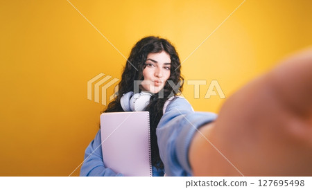 Brunette student taking selfie holding notebook and wearing headphones Brunette student taking selfie holding notebook and wearing headphones 127695498