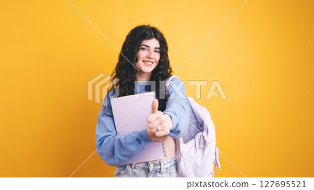 Smiling student showing thumb up holding notebook and backpack on yellow background 127695521