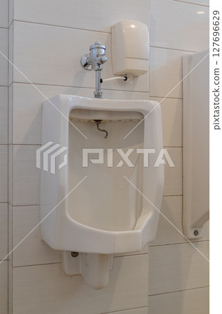 White ceramic urinal with flush valve mounted on tiled wall in public restroom 127696629