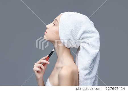 Young caucasian female applying makeup with brush on neck wrapped in towel Young caucasian female applying makeup with brush on neck wrapped in towel 127697414