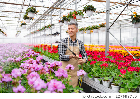 A smiling gardener waters flowers in a vibrant greenhouse, surrounded by colorful blooms, creating a scene of horticultural beauty. A smiling gardener waters flowers in a vibrant greenhouse, surrounded by colorful blooms, creating a scene of horticultural beauty. 127698719