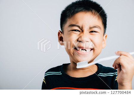 An Asian boy missing an upper milk tooth smiles while holding a toothbrush emphasizing dental care and oral hygiene routine. Isolated on white promoting healthy habits. Children dentist routine An Asian boy missing an upper milk tooth smiles while holding a toothbrush emphasizing dental care and oral hygiene routine. Isolated on white promoting healthy habits. Children dentist routine 127700484