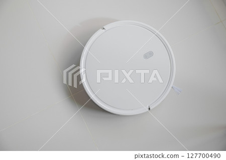 White automatic floor cleaner with side brush on tile flooring. Top view of robot vacuum cleaner removing dust, showcasing modern smart home cleaning technology for spotless and efficient maintenance 127700490