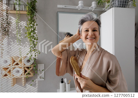 Happy senior woman grooming hair with wooden brush in modern bathroom with natural decor Happy senior woman grooming hair with wooden brush in modern bathroom with natural decor 127700512