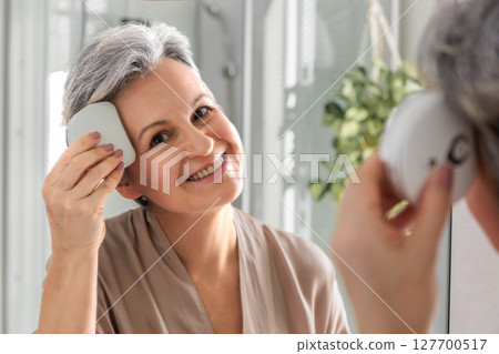 miling mature woman using facial massage device in front of bathroom mirror at home 127700517