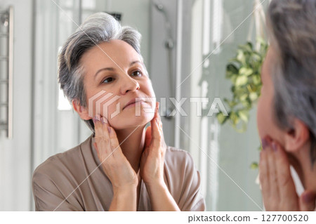 Mature woman examining her face in mirror during morning skincare routine at home Mature woman examining her face in mirror during morning skincare routine at home 127700520