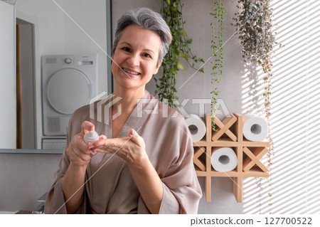 Cheerful mature woman applying skincare product in bathroom with natural morning light 127700522