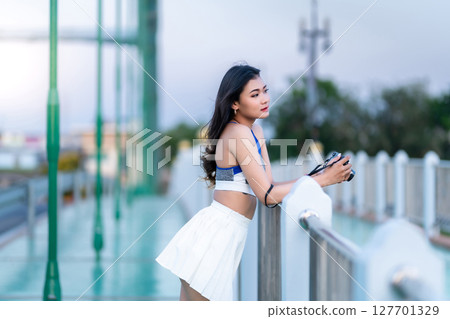 Portrait of beautiful athletic young asian woman in fashionable sportswear standing and with a holding the camera on Route walking jogging or ride bicycle and exercise on the bridge. Portrait of beautiful athletic young asian woman in fashionable sportswear standing and with a holding the camera on Route walking jogging or ride bicycle and exercise on the bridge. 127701329