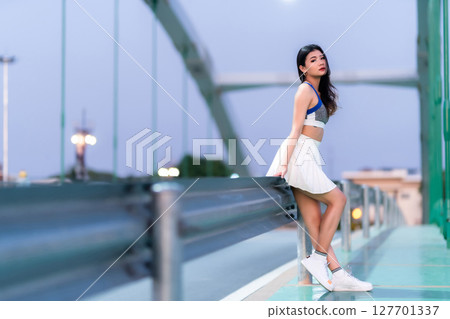 Portrait of beautiful athletic young asian woman in fashionable sportswear standing and having rest after exercise training on Route walking jogging or ride bicycle and exercise on the bridge. 127701337