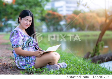 Happy Relaxing Portrait of freelancer asian woman Wear purple dress while working holding diary book writing note while sitting on green grass lawn beside a reservoir at the city park outdoors. 127701706