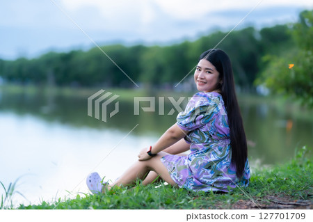 Happy Relaxing Portrait asian woman Wear purple dress wearing smartwatch while sitting on green grass lawn beside a reservoir at the city park outdoors. Happy Relaxing Portrait asian woman Wear purple dress wearing smartwatch while sitting on green grass lawn beside a reservoir at the city park outdoors. 127701709