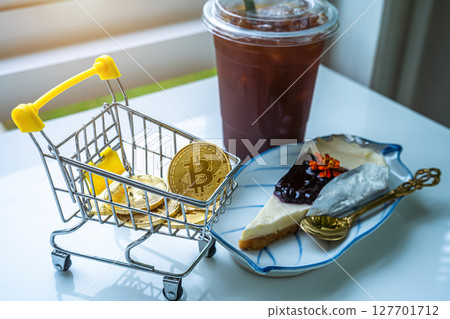 Close-up of Golden bitcoins or cryptocurrency coin or symbols in shopping cart and Americano iced coffee or black and Blueberry Cake in white table. Future currency concept. 127701712