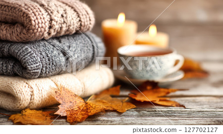 Cozy knitted sweaters stacked with autumn leaves, candles, and a warm cup of tea, creating a serene and inviting atmosphere for fall 127702127