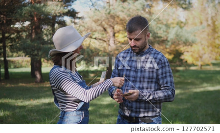Two friends stand in a green park exchanging plants while enjoying a sunny day. The bright foliage creates a vibrant atmosphere for their conversation. Two friends stand in a green park exchanging plants while enjoying a sunny day. The bright foliage creates a vibrant atmosphere for their conversation. 127702571