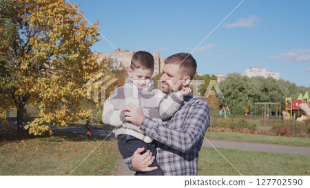 In a bright autumn park, a father gently holds his son, surrounded by colorful foliage and a playground, enjoying quality time on a sunny day. In a bright autumn park, a father gently holds his son, surrounded by colorful foliage and a playground, enjoying quality time on a sunny day. 127702590