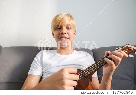 Caucasian Boy Playing Ukulele while Sitting on Sofa, Looking into Camera. Young Musician Learning to Play Instrument at Home. Caucasian Boy Playing Ukulele while Sitting on Sofa, Looking into Camera. Young Musician Learning to Play Instrument at Home. 127702849