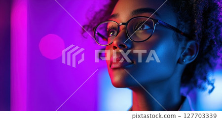 Young woman with glasses gazes thoughtfully under vibrant neon lights in a modern indoor setting during the evening 127703339