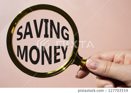 Saving Money symbol. Concept words Saving Money on magnifying glass. Businessman hand. Beautiful pink background. Business and Saving Money concept. Copy space. Saving Money symbol. Concept words Saving Money on magnifying glass. Businessman hand. Beautiful pink background. Business and Saving Money concept. Copy space. 127703559