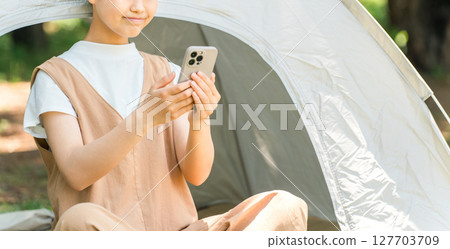 一個女孩在露營地的帳篷前看著她的智慧型手機 一個女孩在露營地的帳篷前看著她的智慧型手機 127703709
