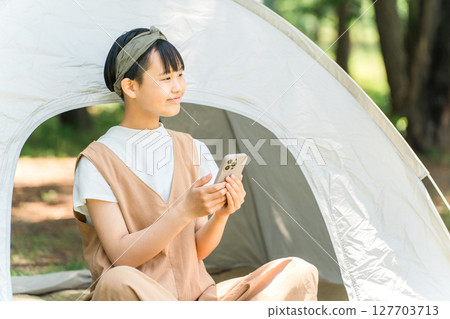 A girl looking at her smartphone in front of a tent at a campsite 127703713