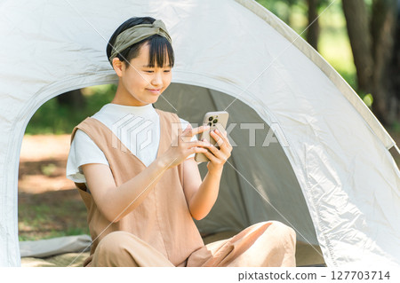 A girl looking at her smartphone in front of a tent at a campsite 127703714