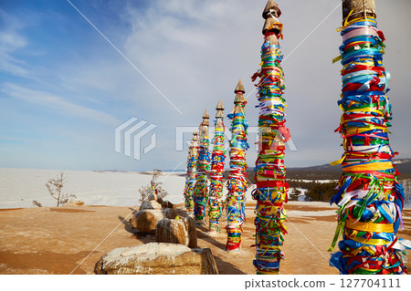 Lake Baikal in winter. Olkhon Island. Wooden ritual pillars with colorful ribbons on cape Burhan or Shamanka rock Lake Baikal in winter. Olkhon Island. Wooden ritual pillars with colorful ribbons on cape Burhan or Shamanka rock 127704111