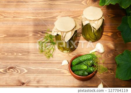 Pickled cucumbers in jars, fresh cucumbers on bowl, dill, garlic 127704119