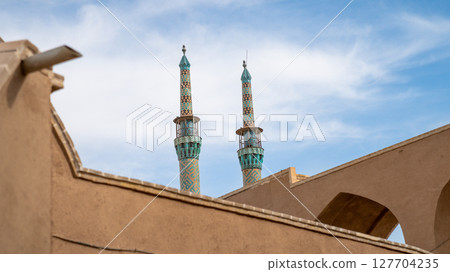 Intricately tiled minarets of Jameh Mosque of Yazd, rising above traditional mud-brick buildings under a sunny sky in Yazd Intricately tiled minarets of Jameh Mosque of Yazd, rising above traditional mud-brick buildings under a sunny sky in Yazd 127704235