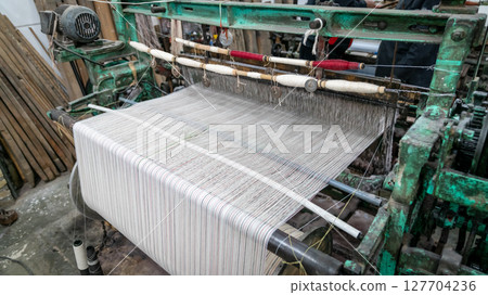 Close-up of a vintage textile weaving loom in operation, showing threads forming a striped fabric, highlighting traditional production, Iran Close-up of a vintage textile weaving loom in operation, showing threads forming a striped fabric, highlighting traditional production, Iran 127704236