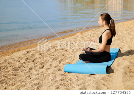 A young girl is sitting in the lotus position with her eyes closed on a sandy beach. Time for yoga. Meditation and rest 127704535