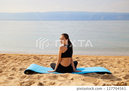 A sporty girl is sitting on a yoga mat on a sandy beach on the shore of a lake. Yoga time. 127704561