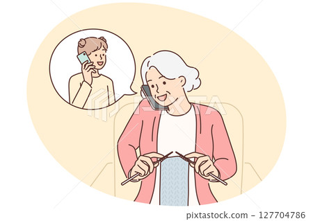 Grandmother makes phone call to granddaughter and knits scarf sitting in cozy chair 127704786