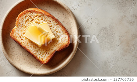 Slice of toasted bread with two pieces of butter served on a ceramic plate over a light textured surface. Simple and rustic breakfast. Warm and cozy. Slice of toasted bread with two pieces of butter served on a ceramic plate over a light textured surface. Simple and rustic breakfast. Warm and cozy. 127705736