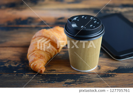 Coffee cup with black lid beside a flaky croissant and tablet on rustic wooden table, creating a cozy breakfast scene for morning enjoyment 127705761