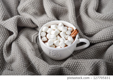 Hot chocolate in a white cup topped with marshmallows and a cinnamon stick, placed on a cozy gray knitted blanket for a warm atmosphere 127705831