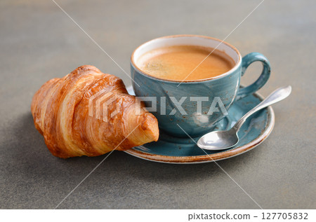 Freshly brewed coffee in a blue ceramic cup beside a flaky croissant on a rustic plate, creating a cozy breakfast scene with inviting ambiance 127705832