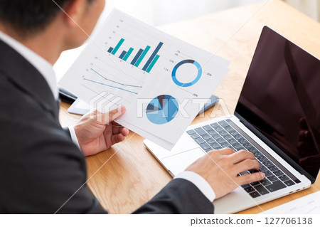 A middle-aged male businessman in a suit working on a computer while looking at graphs at his desk 127706138