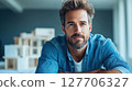 Young man with a beard poses confidently in a modern workspace, showcasing creativity and approachability while surrounded by architectural models 127706327