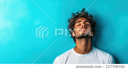Young man with curly hair relaxes against a blue wall, eyes closed, in a peaceful moment captured in a bright and vibrant atmosphere 127706382