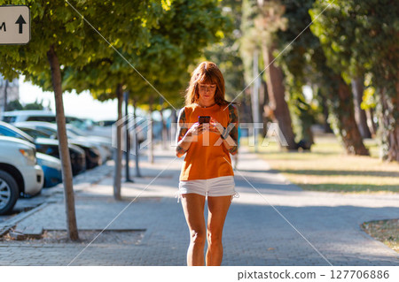Young Caucasian tanned woman with tattoo using cellphone in her hands and walking down the street. Concept of online communication Young Caucasian tanned woman with tattoo using cellphone in her hands and walking down the street. Concept of online communication 127706886